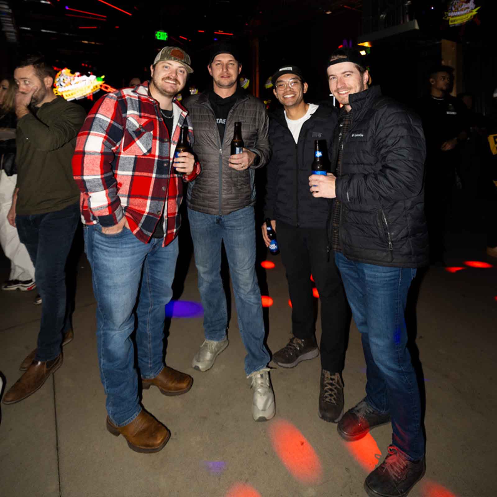 Group of four men enjoying drinks together in a lively bar setting with colorful lights.