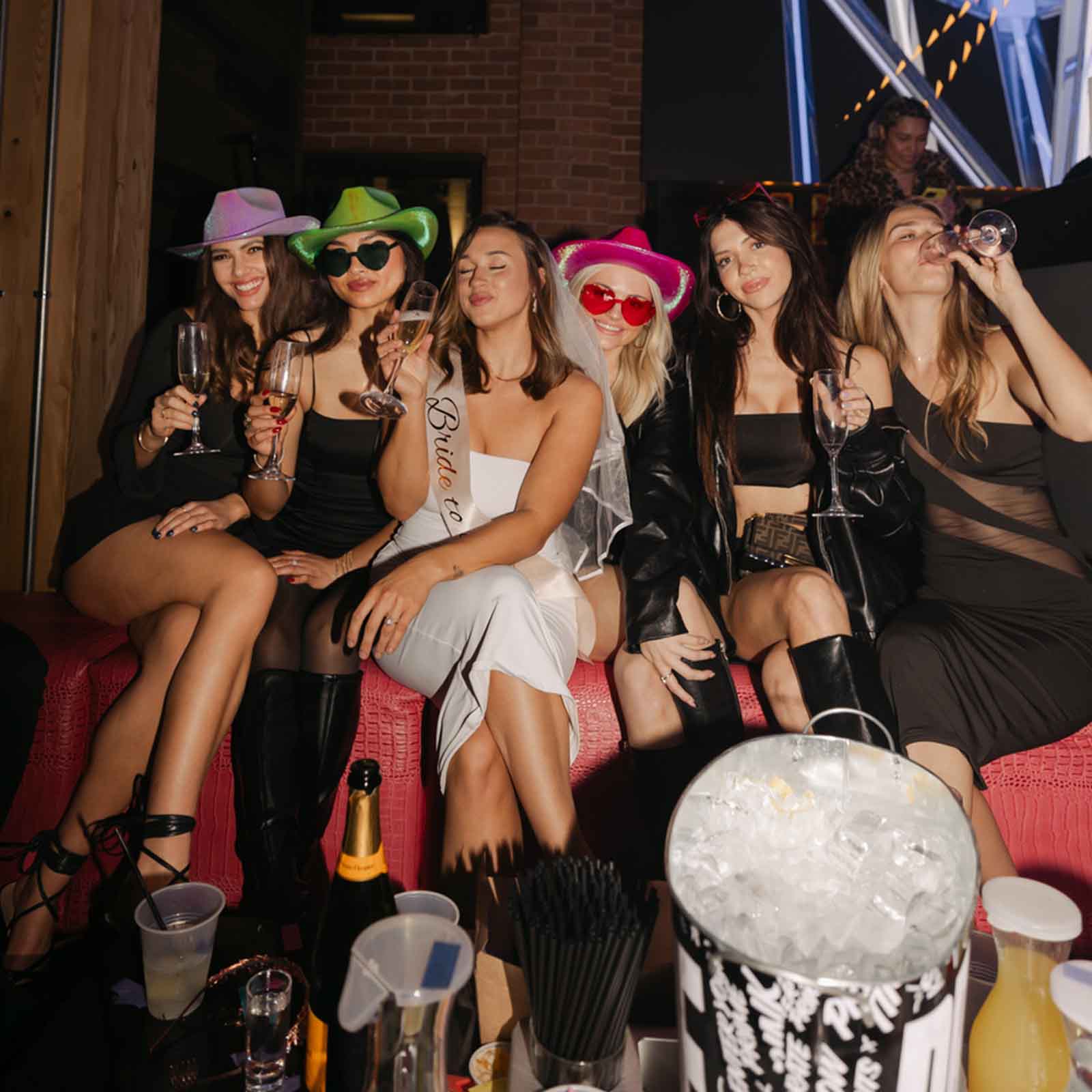 Group of women celebrating with drinks, wearing fun hats and glasses at a lively party.