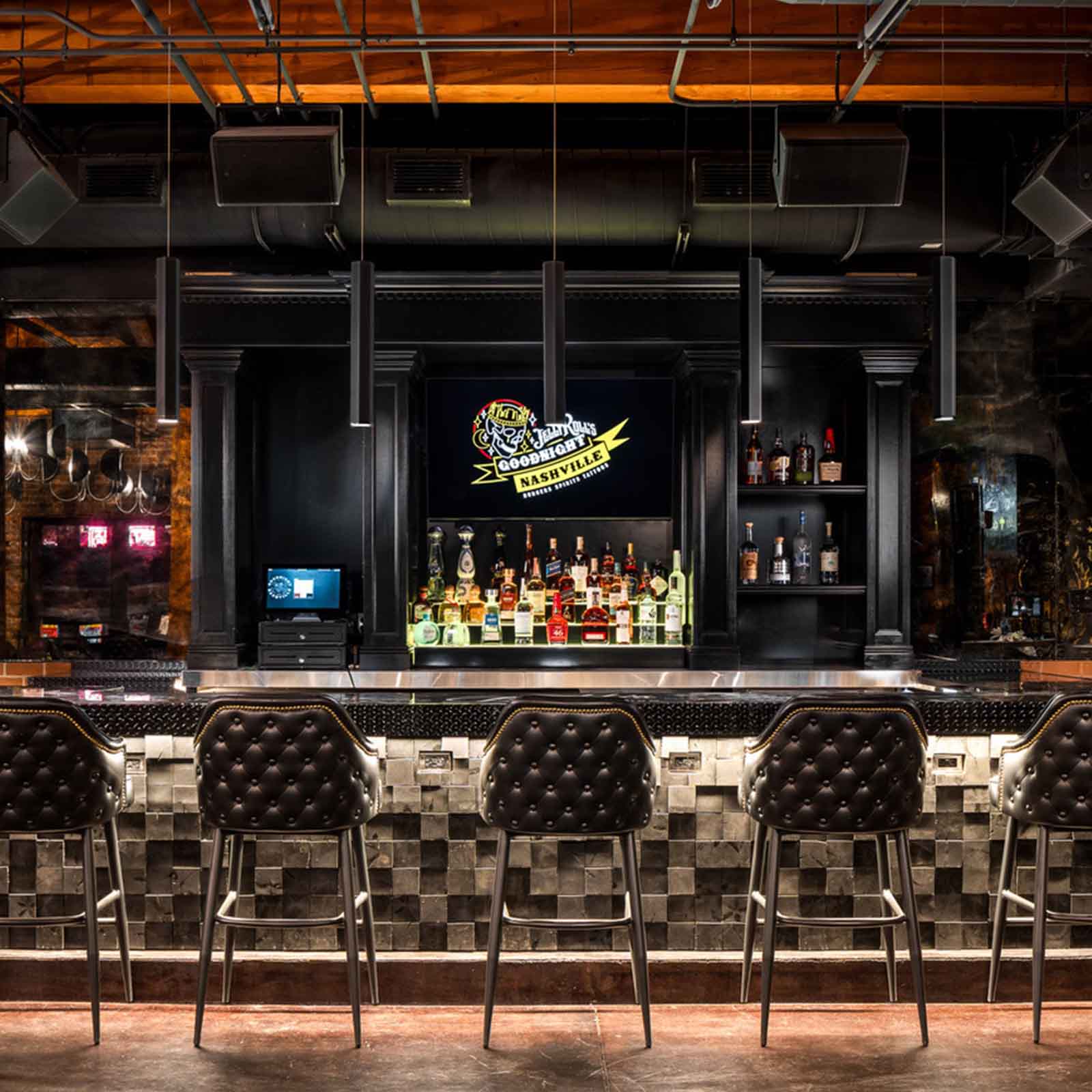 A stylish bar with a black and silver tiled counter, tufted barstools, and a well-stocked liquor display under dim lighting