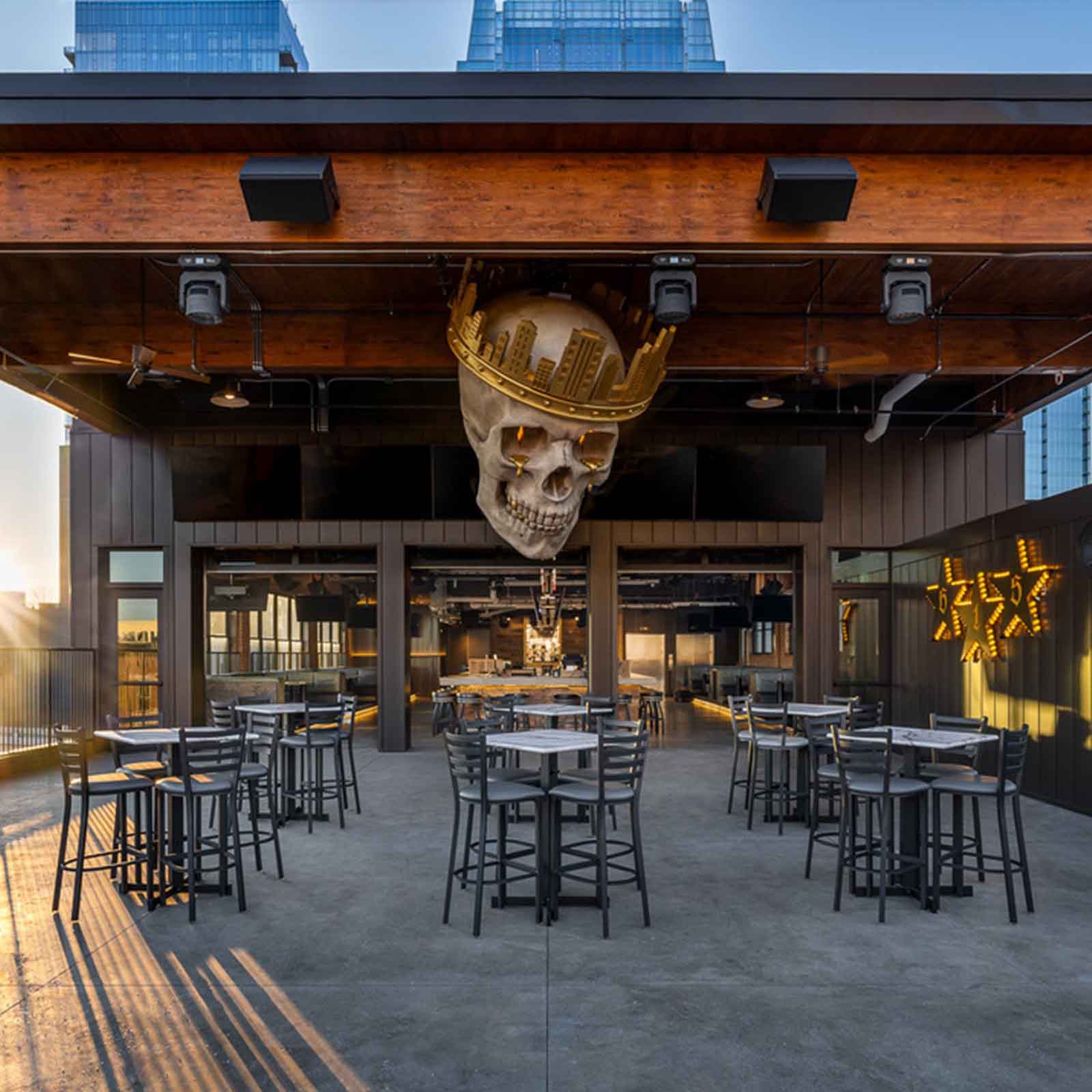 A rooftop bar with industrial-style decor, high-top tables, and a large skull sculpture wearing a golden crown with a city skyline design, suspended from the ceiling.