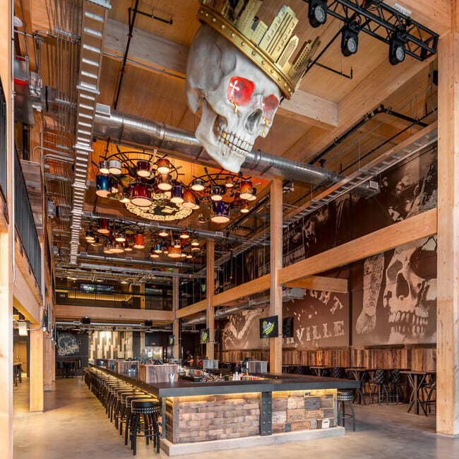 A bar inside a large venue with a dramatic overhead sculpture of a skull wearing a crown. The space features industrial design elements with exposed pipes and a modern bar area.