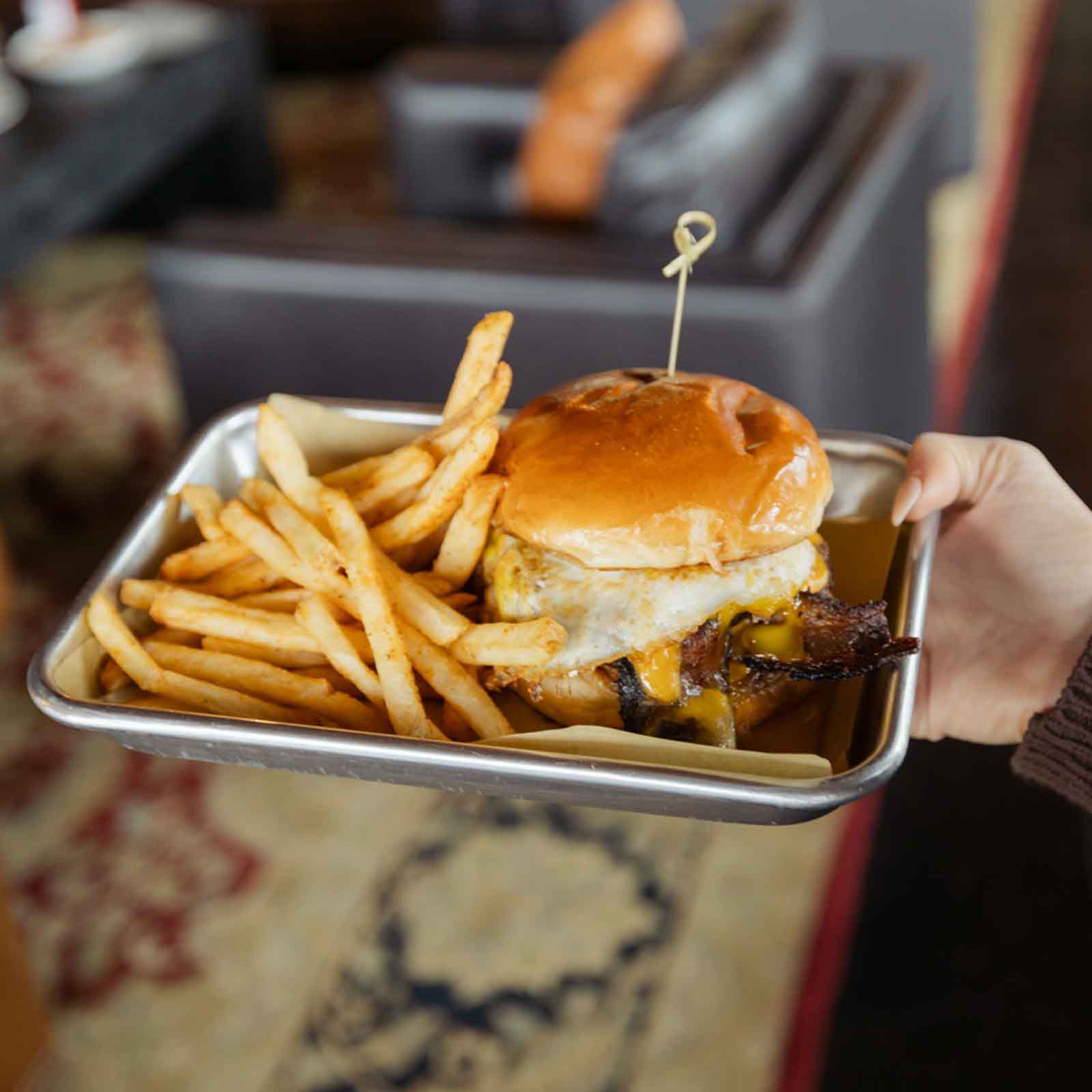 A close-up of a burger with cheese and a fried egg on top, served with a side of fries in a metal tray.