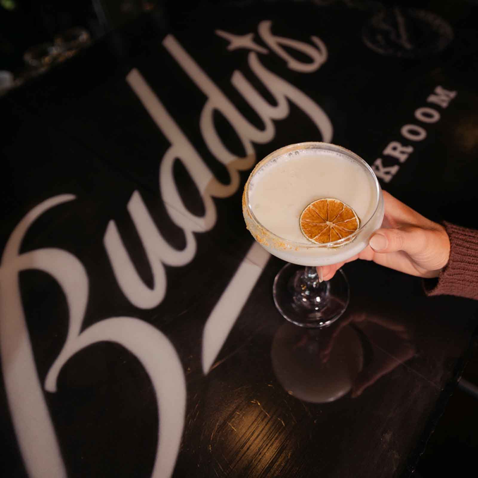 A cocktail in a coupe glass with a dried citrus garnish, placed on a bar counter with the word "Buddy