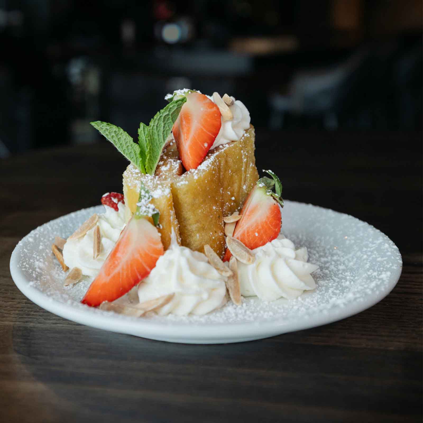 A dessert plate with a portion of French toast, topped with fresh strawberries, whipped cream, mint leaves, and slivered almonds. The dish is dusted with powdered sugar for a finishing touch.