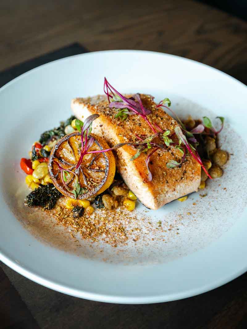 A plate of grilled fish fillet, garnished with microgreens, a charred lemon slice, and a dusting of seasoning.