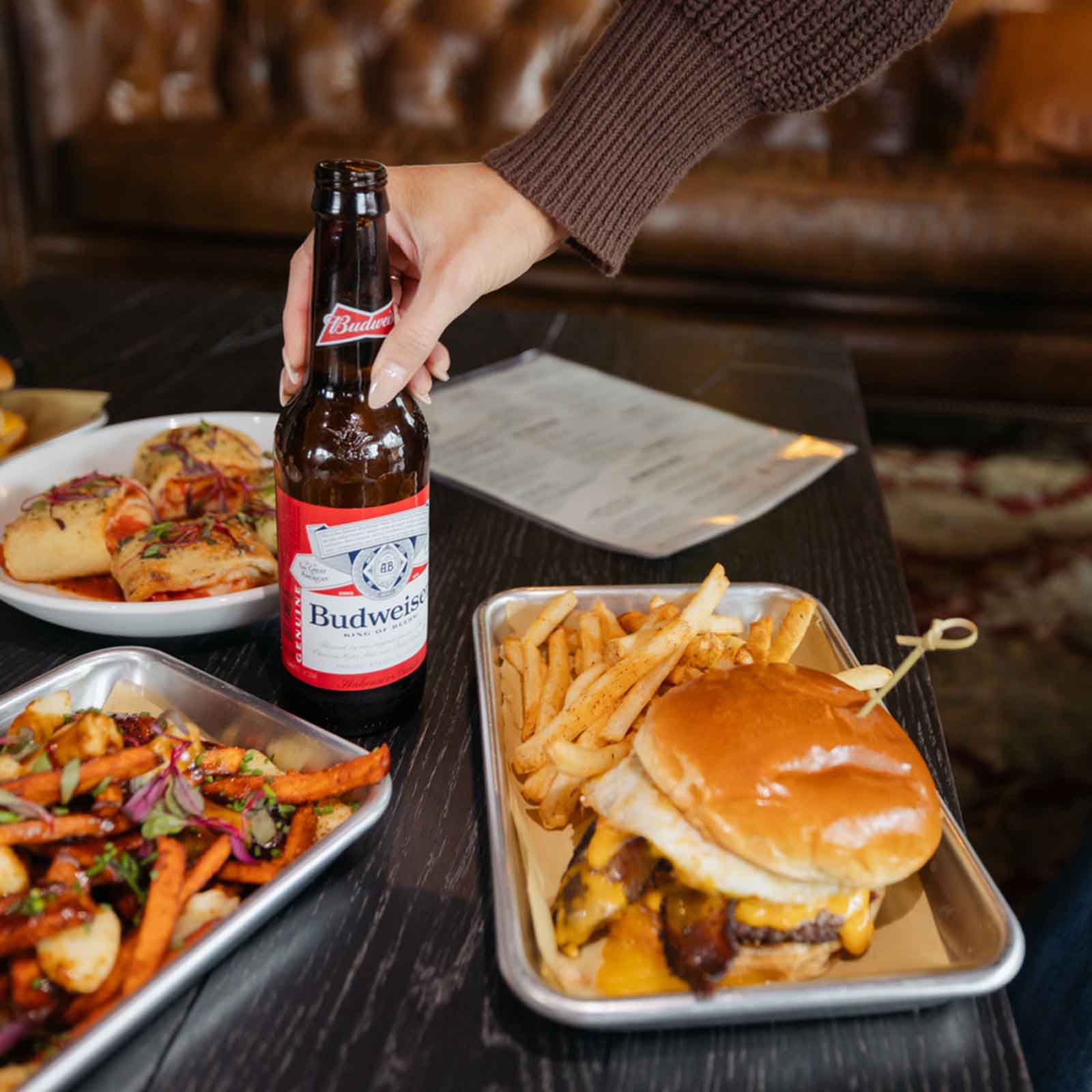 A bottle of Budweiser beer next to a plate with a burger, fries, and a dish of seasoned sweet potato fries with toppings.