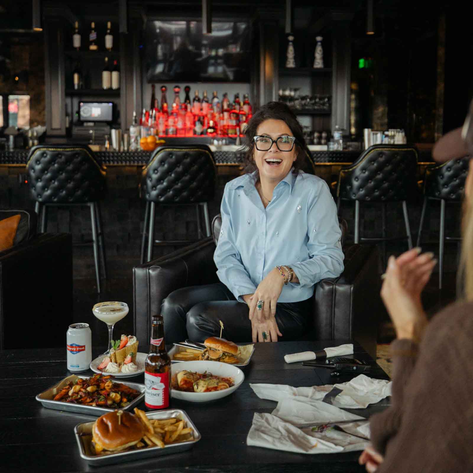 A woman smiling and sitting at a table with several food and drinks. The table includes a burger with fries, a bowl of pasta, and beverages including a beer and a cocktail. The background features a bar with illuminated bottles.