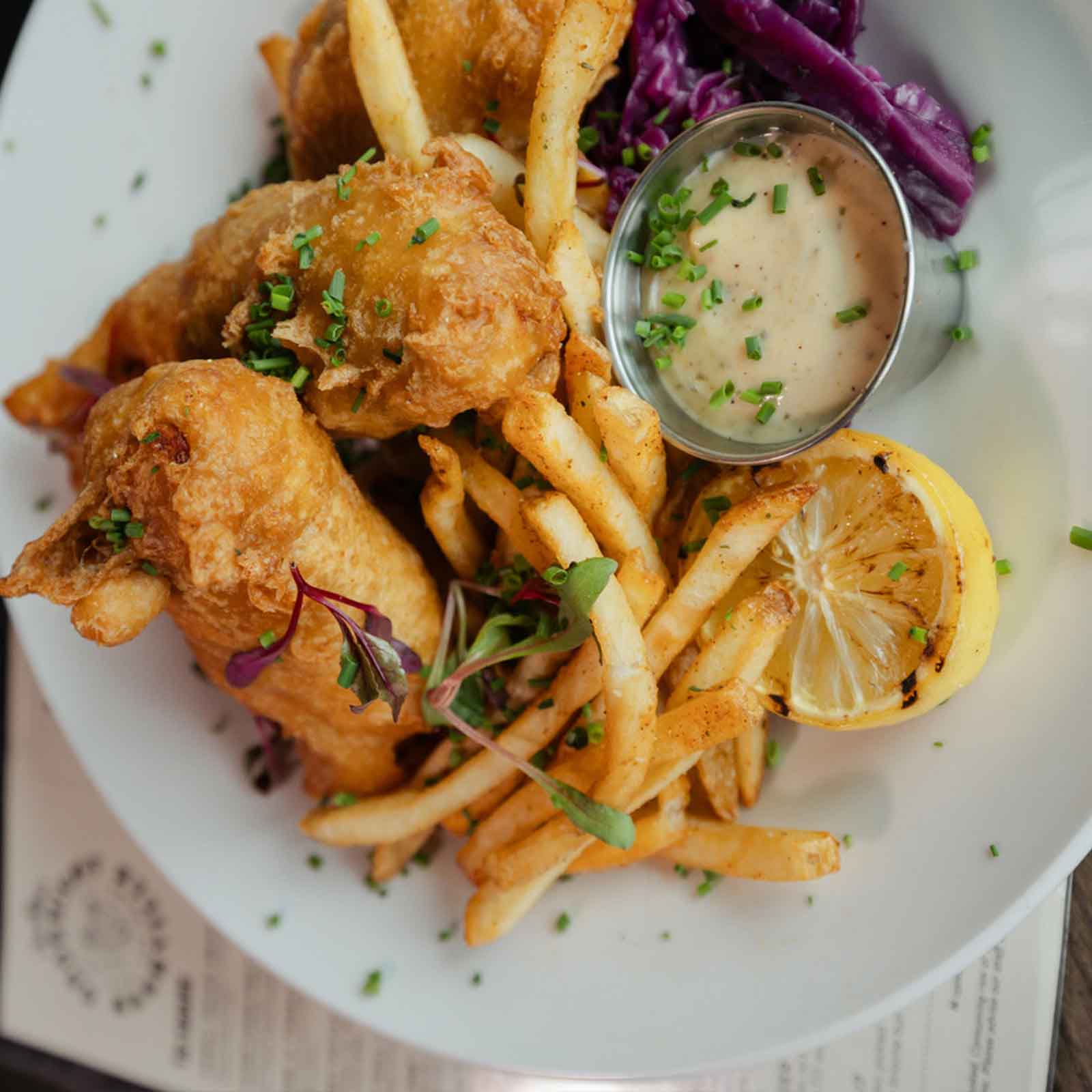 A plate of crispy fried fish, paired with golden French fries, coleslaw, grilled lemon wedges, and a side of dipping sauce, garnished with chopped chives.