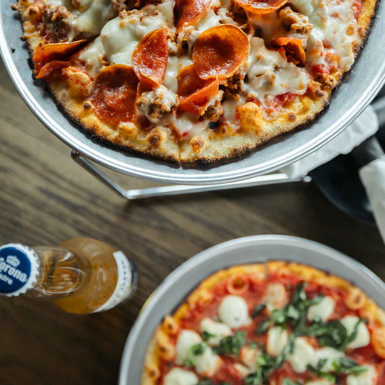 The image shows two pizzas, one with pepperoni and sausage on top of melted cheese, and the other topped with mozzarella and fresh basil leaves. A bottle of Corona beer is placed near the pizzas on the table.