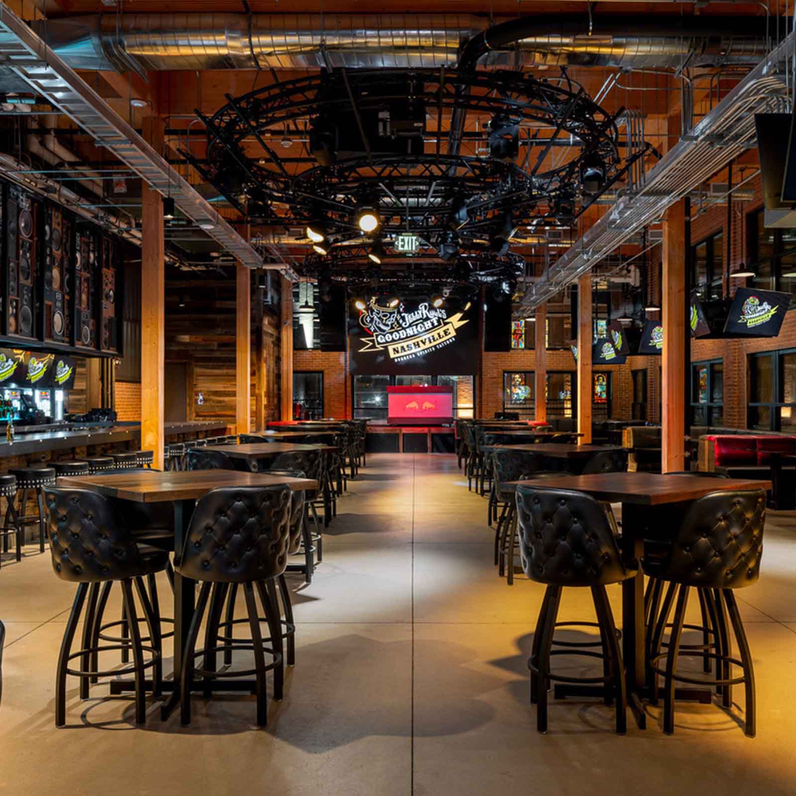 A lively venue with high-top tables and leather chairs, featuring a large circular chandelier with lights and speakers above, creating an industrial and vibrant atmosphere. The venue has a bar and colorful signage with the name 