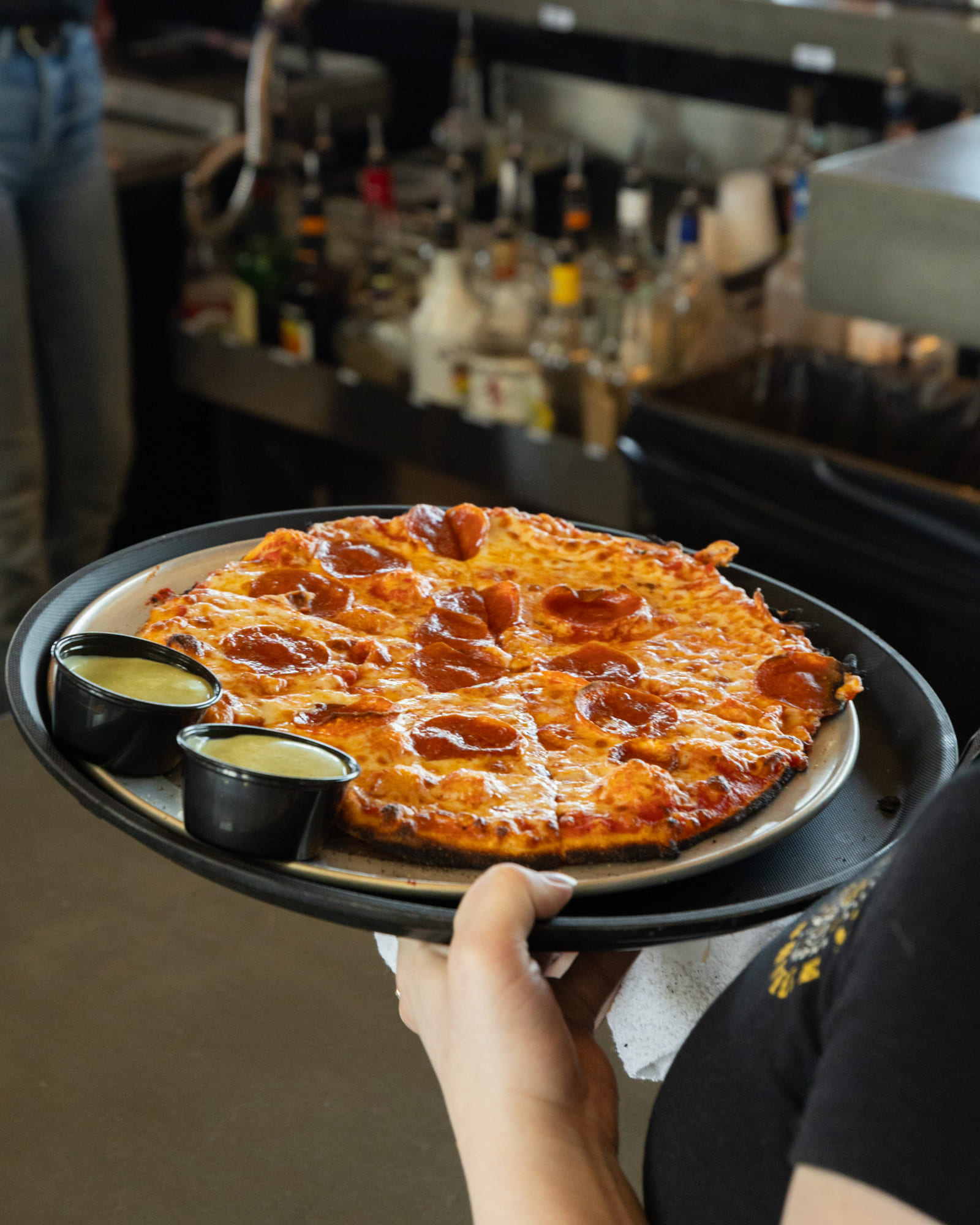 Pizza with melted cheese and pepperoni toppings, served on a black plate with two small dipping cups