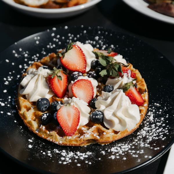 Waffle topped with whipped cream, fresh strawberries, blueberries, and powdered sugar on a black plate.