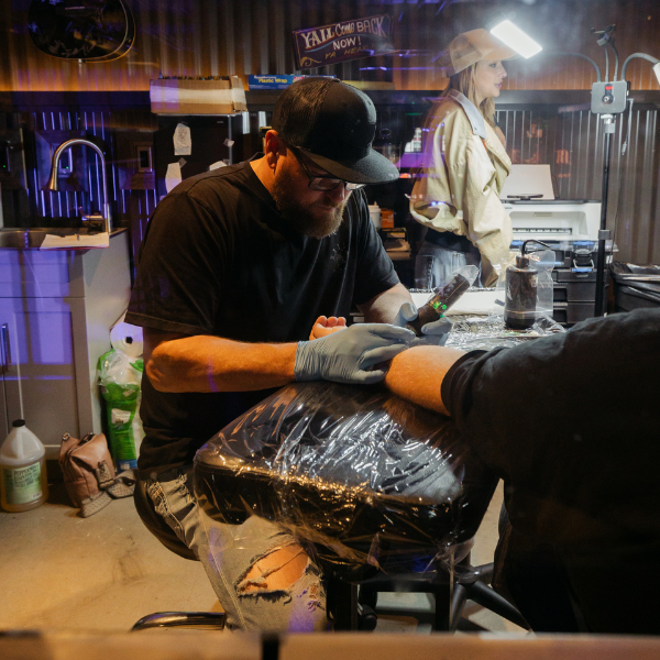 Tattoo artist focused on inking a client’s arm inside a studio with dim lighting and a bystander nearby.