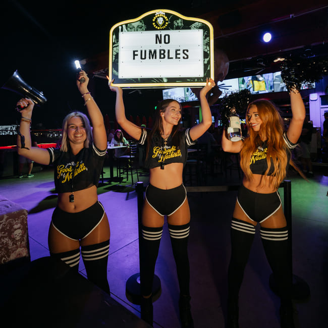 Three women wearing black DawgHouse uniforms celebrate inside a nightclub while holding a glowing sign that reads “No Fumbles.”