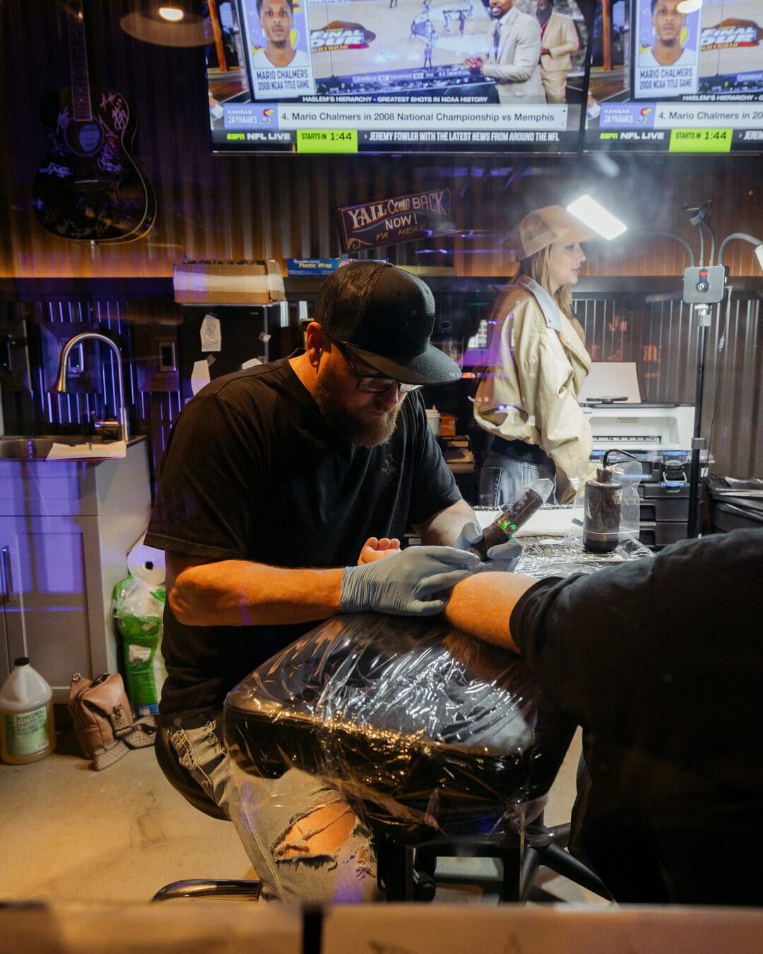Tattoo artist at work in a country studio, focusing on detailed ink design while a TV shows sports highlights in the background.