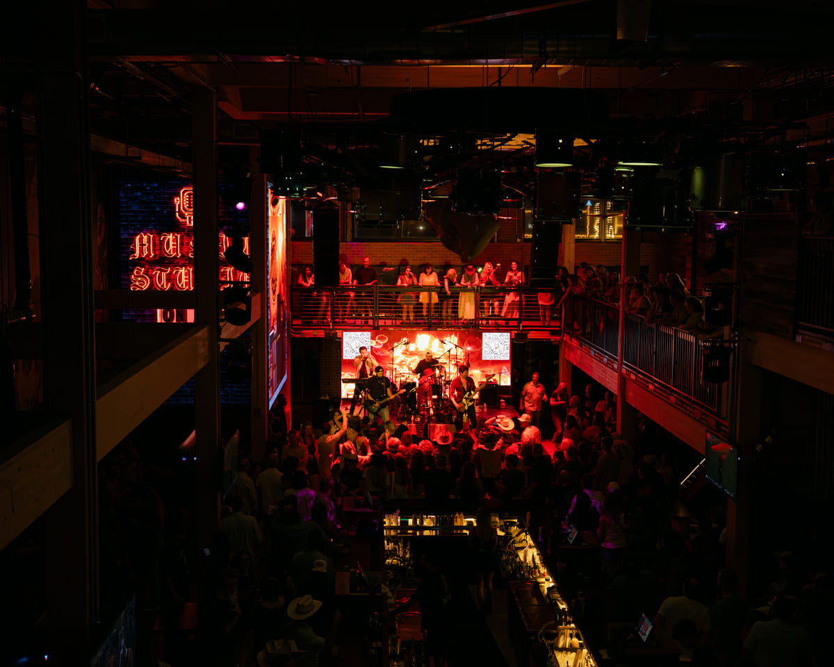 Crowd enjoying a live music performance in a dark, vibrant venue with red stage lighting and upper balcony seating.