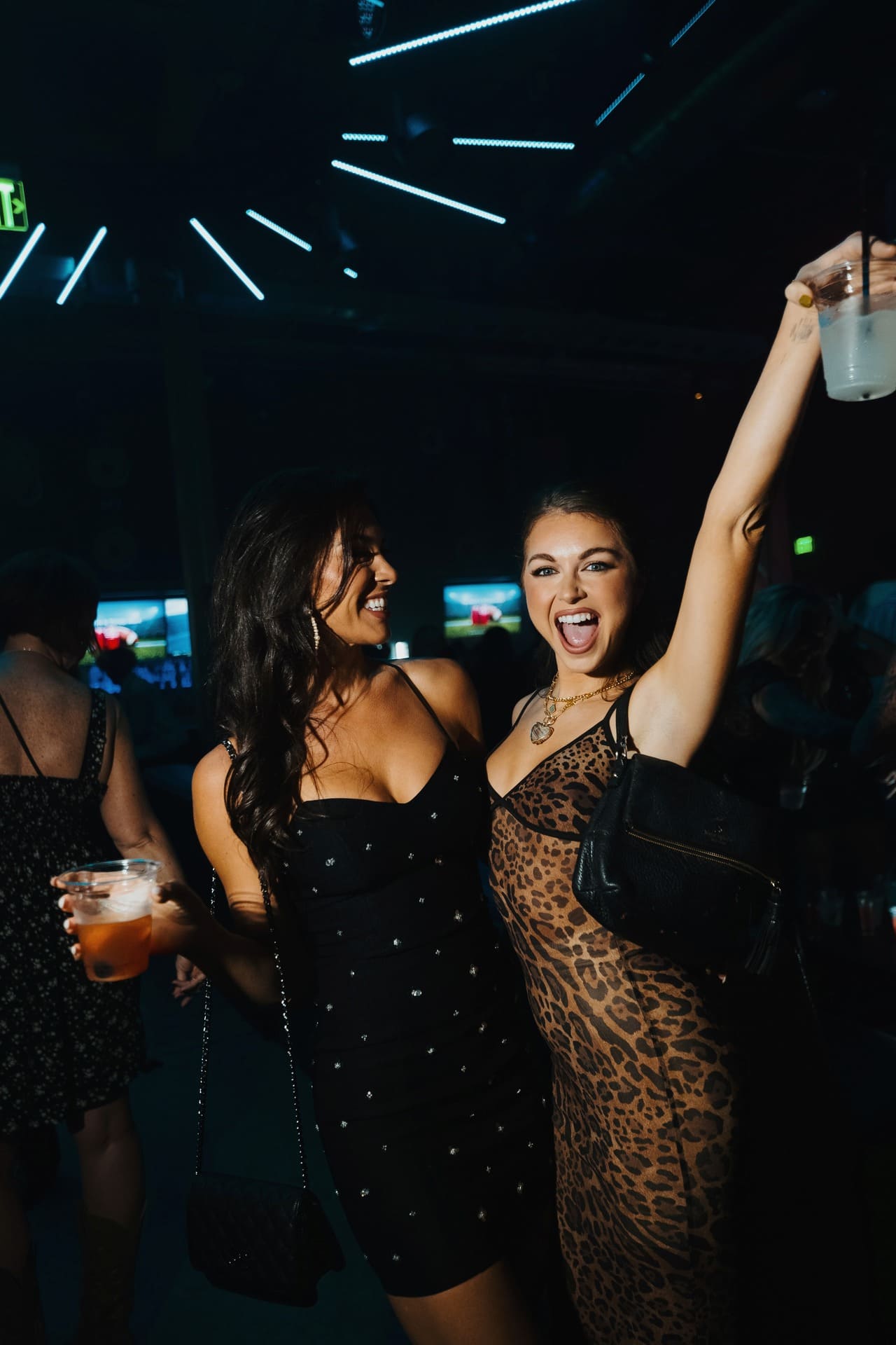 Two women dancing and celebrating at a dimly lit nightclub; one holds a drink while the other raises her arm excitedly under vibrant ceiling lights.