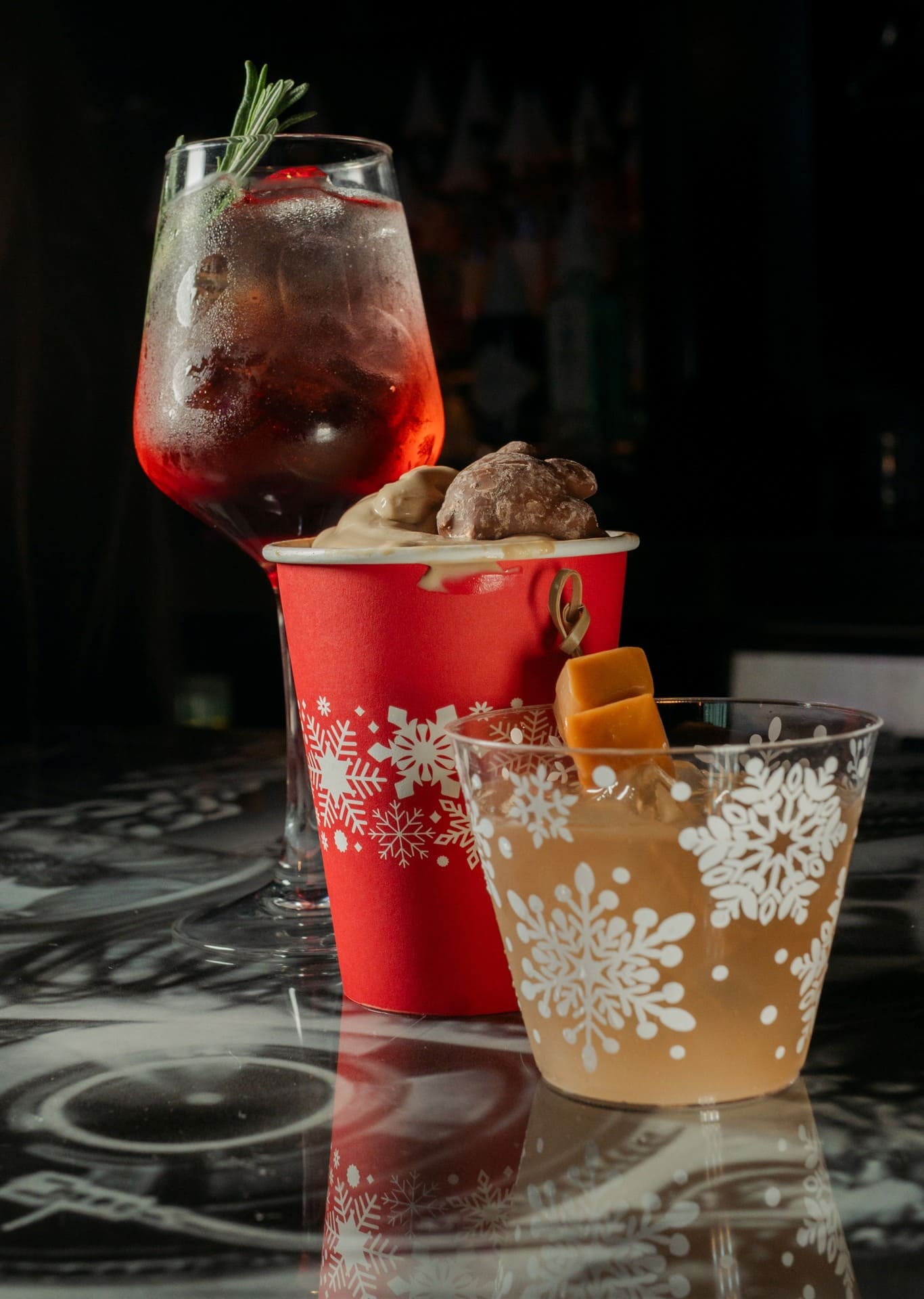 Three festive cocktails sit on a reflective bar counter: a red holiday cup overflowing with creamy dessert and chocolate truffles, a clear glass with a caramel cube garnish, and a tall iced cocktail with rosemary and berries, all decorated with snowflake patterns.