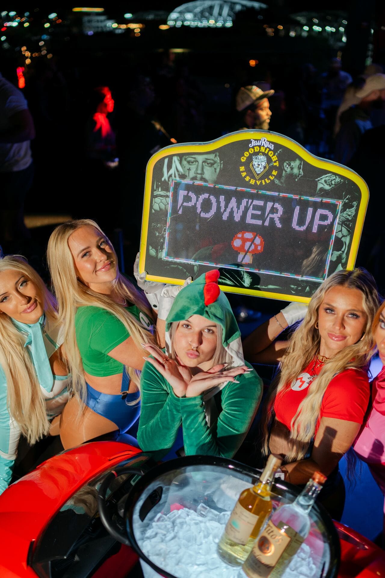 Five women in playful Mario-themed costumes pose at a nightclub, holding a bright “Power Up” sign above a red toy car filled with iced bottles.