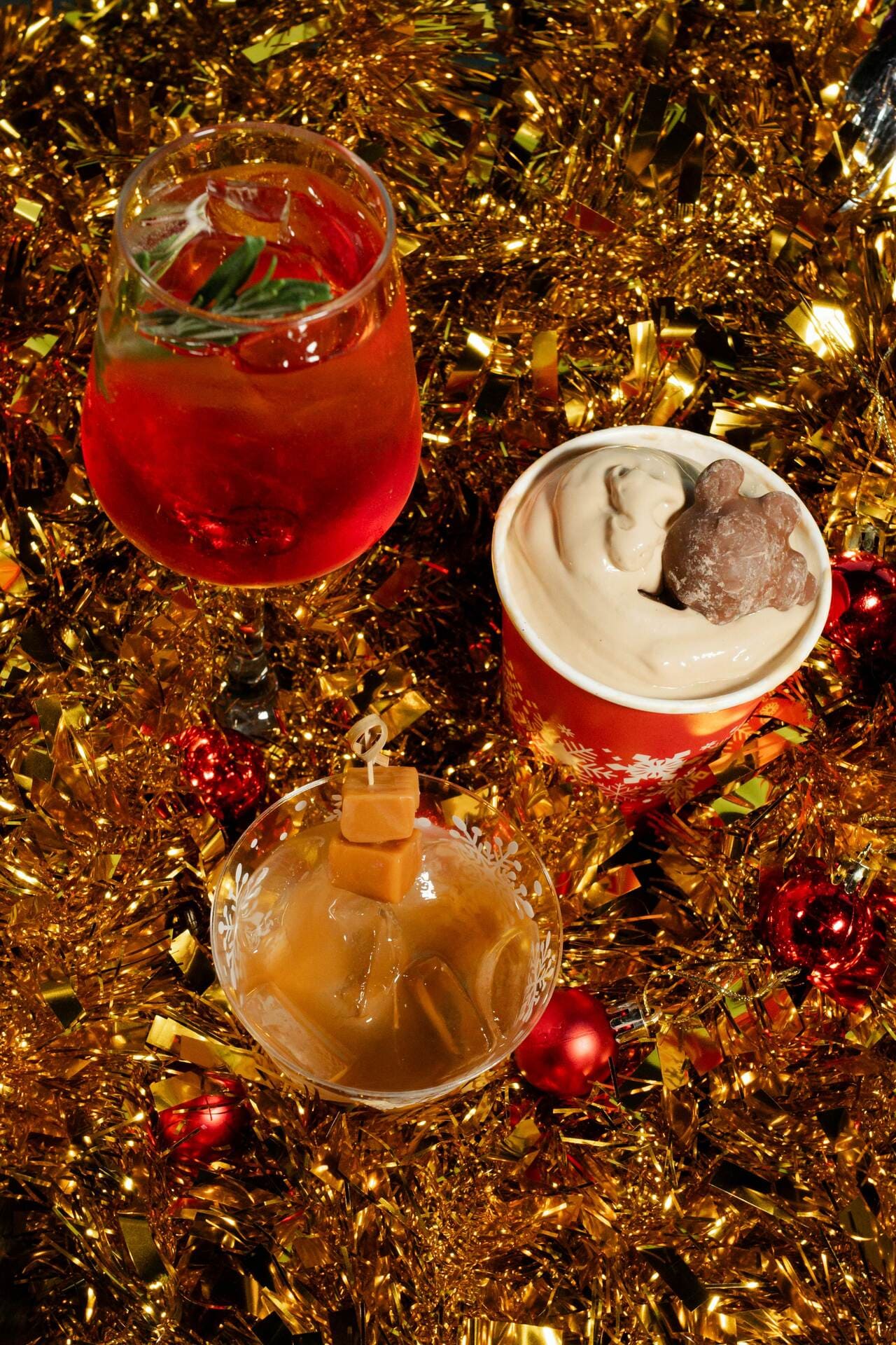 Three festive holiday drinks—one with red spritz and rosemary, one creamy dessert drink with chocolate, and one caramel cocktail—are arranged on a bed of shiny gold tinsel with red ornaments.
