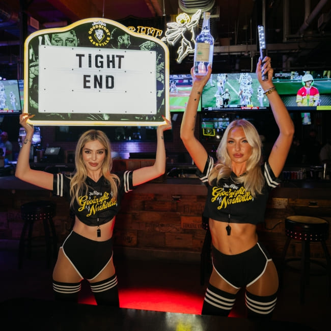 Two women in branded outfits holding a football-themed sign and bottles inside a bar with sports screens