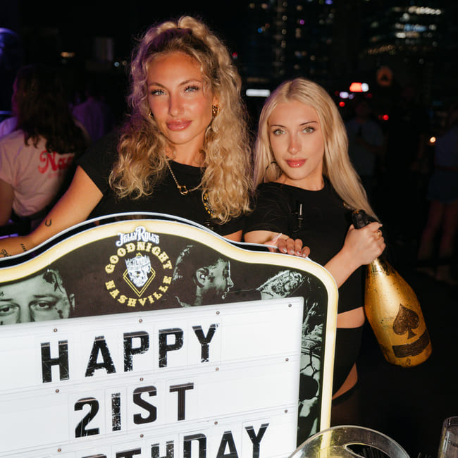 Two women celebrating a 21st birthday at a nightclub holding a champagne bottle and sign