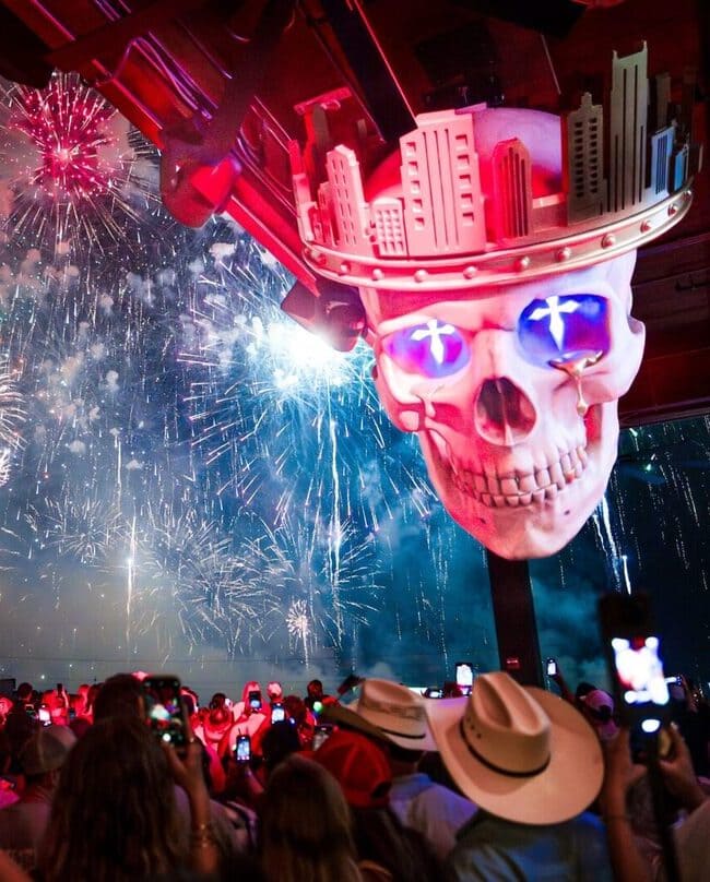 Large illuminated skull art installation above a rooftop crowd watching fireworks during a nighttime celebration.